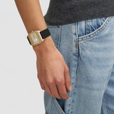 Rückansicht der LIEBESKIND BERLIN Vinyl Vision Damenuhr mit Gehäuse in Gold und schwarzem Lederarmband, getragen am Handgelenk zu Jeans und dunkelgrauem Oberteil.
