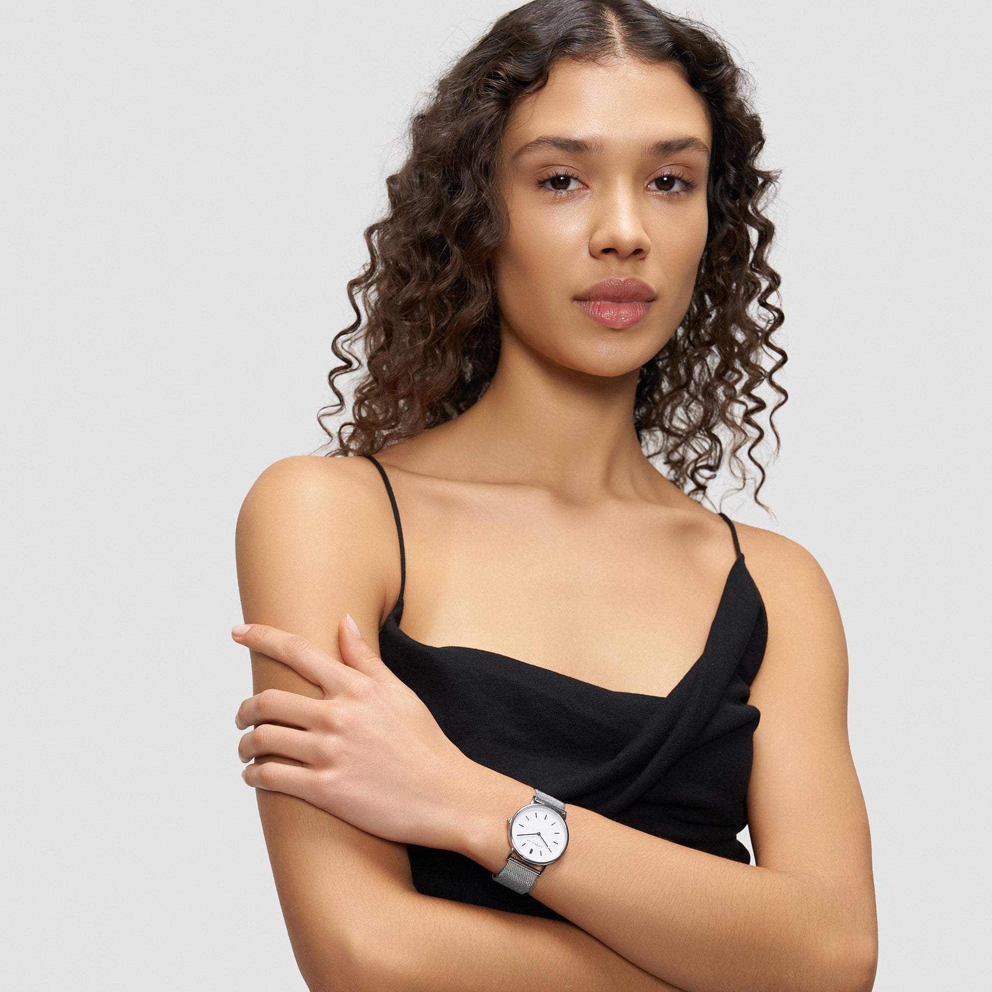 Eine Frau mit lockigem Haar in einem schwarzen Kleid trägt die Liebeskind Berlin Pure Elegance Armbanduhr und posiert vor einem schlichten hellgrauen Hintergrund.