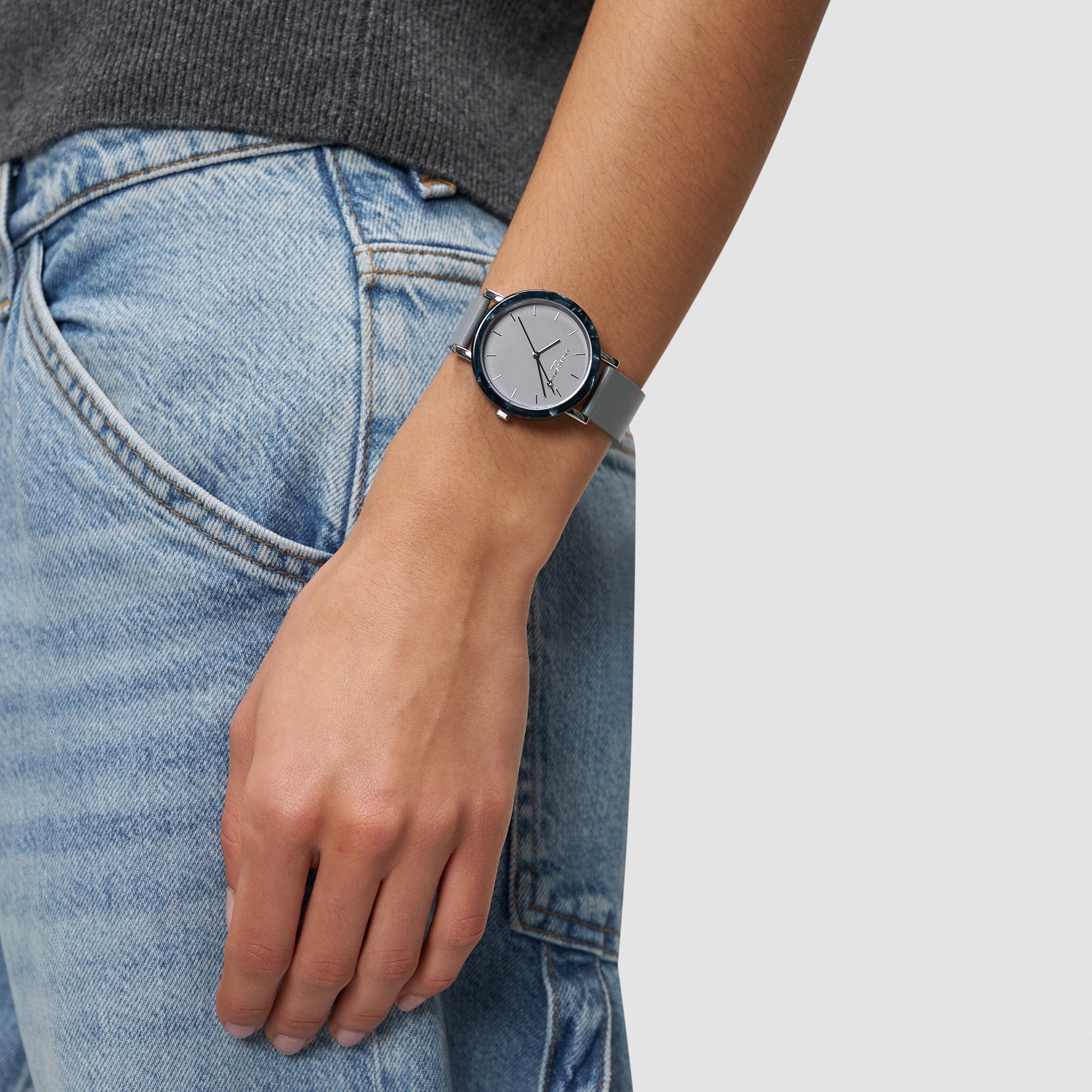 Eine Person trägt die LIEBESKIND BERLIN Armbanduhr - Acetat Chic von Liebeskind Berlin, silberfarben, kombiniert mit blauen Jeans und locker an der Seite gehalten.