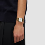 Eine Person in Schwarz trägt die Liebeskind Berlin Armbanduhr - Modern Mesh mit quadratischem, goldenem Gehäuse, weißem Zifferblatt und Edelstahlarmband aus Mesh, den Arm entspannt an der Seite.