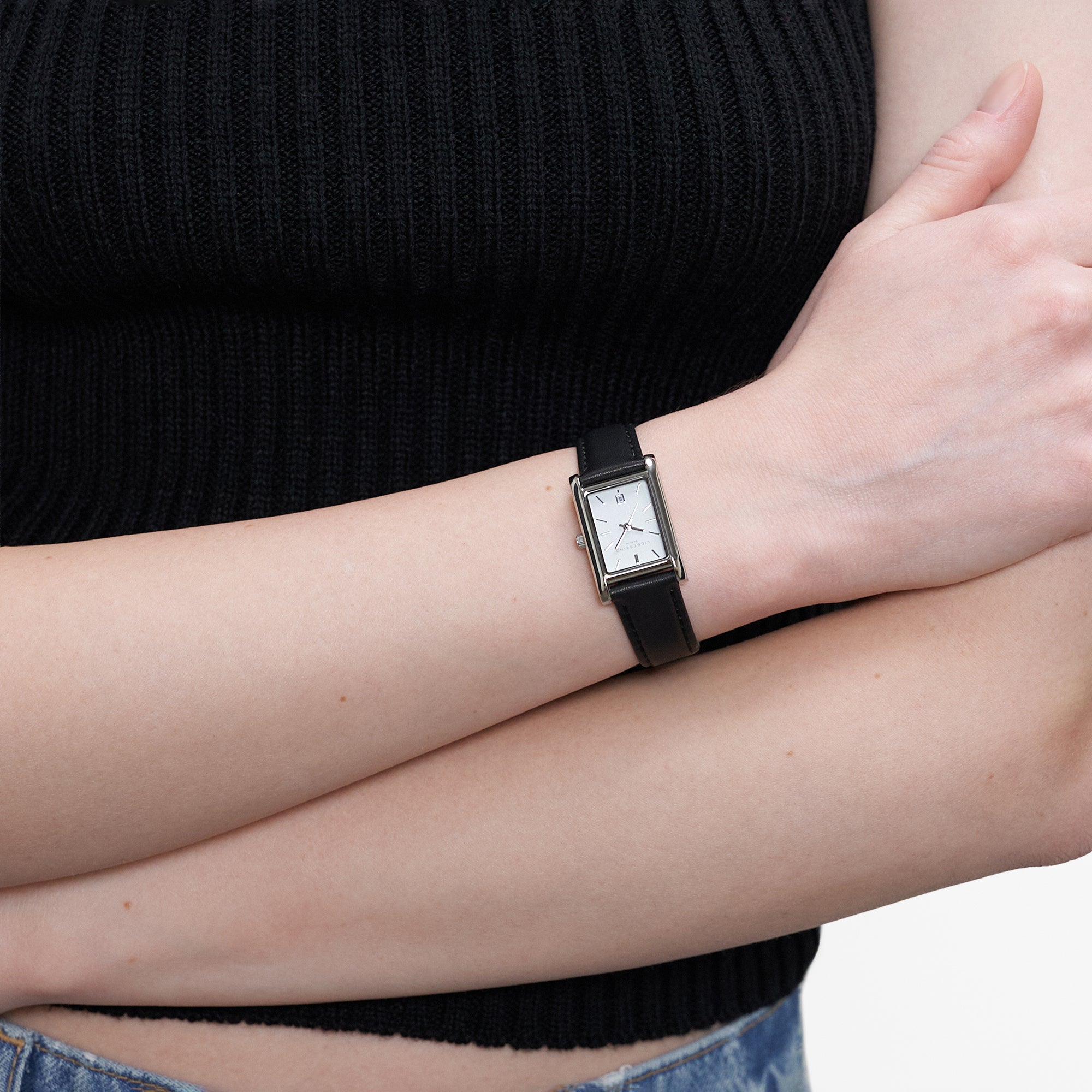Rechteckige LIEBESKIND BERLIN Damenarmbanduhr mit schwarzem Lederarmband und weißem Zifferblatt am Handgelenk getragen.