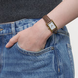 LIEBESKIND BERLIN Damenarmbanduhr mit rechteckigem goldfarbenem Gehäuse und braunem Lederarmband am Handgelenk getragen.