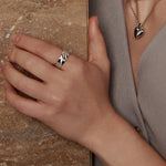 Eine Hand mit einem Silberring ruht auf einer Steinfläche; außerdem ist eine Halskette "Halskette - Schildanhänger" von Ania Haie zu sehen.