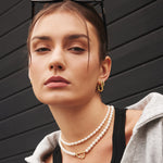Frau mit der ANIA HAIE Halskette - Pearls by Ania Haie, goldenen Ohrringen und Sonnenbrille auf dem Kopf, posiert vor einem dunklen Holzhintergrund.