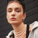 Frau mit neutralem Gesichtsausdruck trägt die ANIA HAIE Halskette - Shell Pearl von Ania Haie, Ohrringe und einen grauen Kapuzenpullover vor einer dunklen Wand.