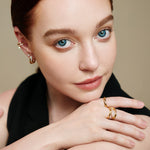 Eine Frau mit blauen Augen, die Goldschmuck und den Ania Haie Ringe - Gold Sparkle Ring 54 von Ania Haie trägt, posiert in einem schwarzen Top mit der Hand am Kinn und schaut in die Kamera.