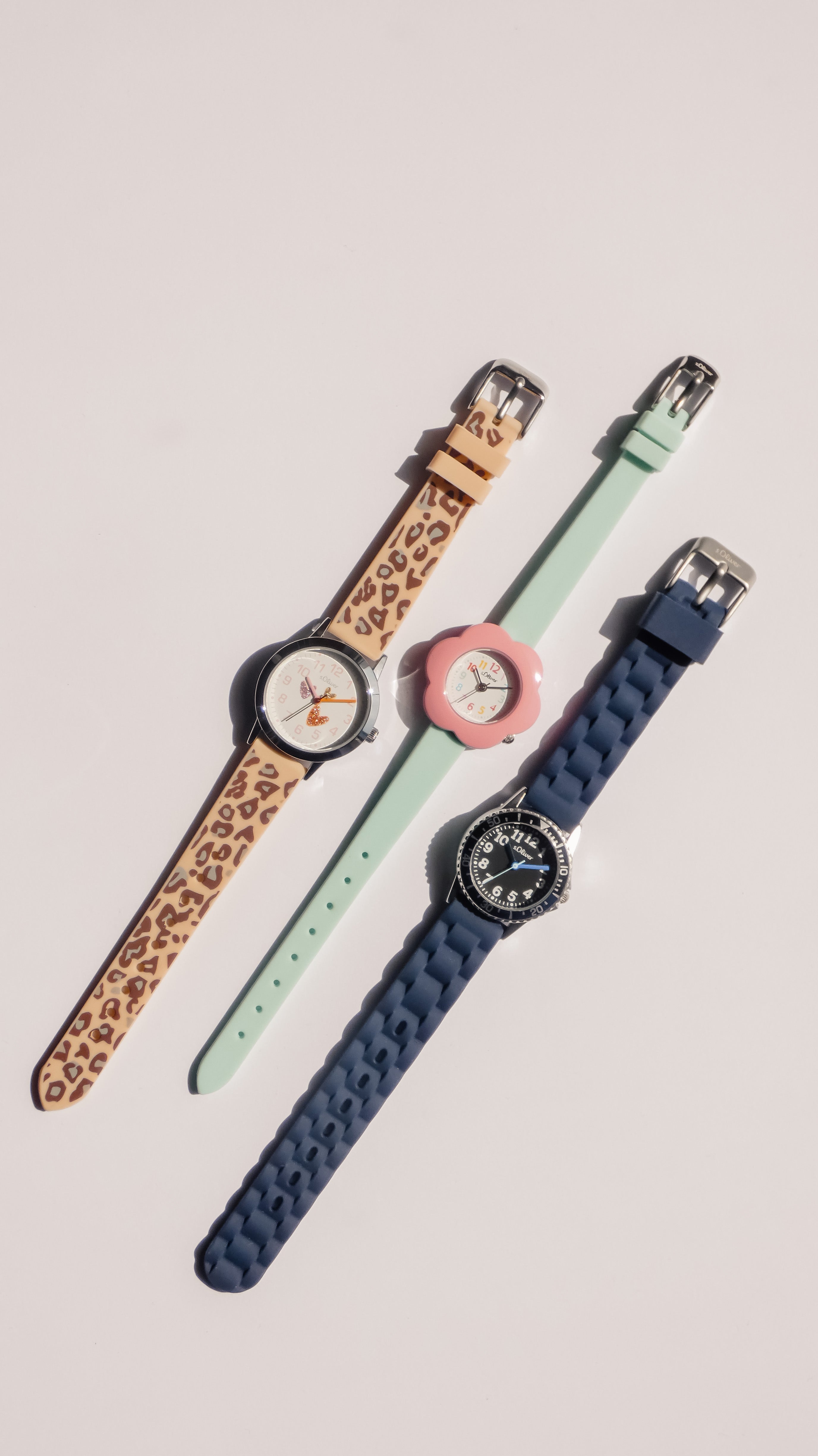 Drei Kinderarmbanduhren mit bunten Armbändern (Leopardenmuster, Mintgrün und Dunkelblau) auf hellem Hintergrund.