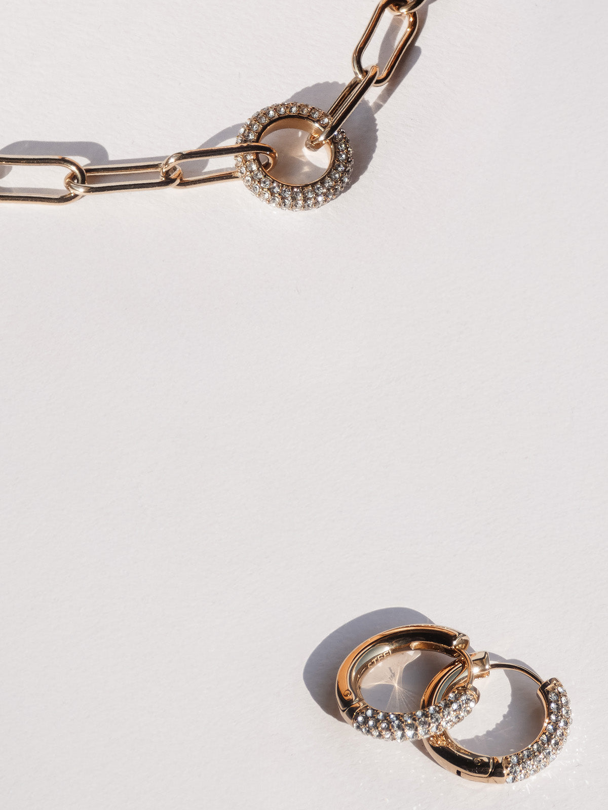 Tamaris Schmuck im Glitter-Donut-Design: goldfarbene Kette und Creolen mit funkelnden, rund gefassten Steinen, modern arrangiert auf hellem Hintergrund mit sanften Lichtreflexen.