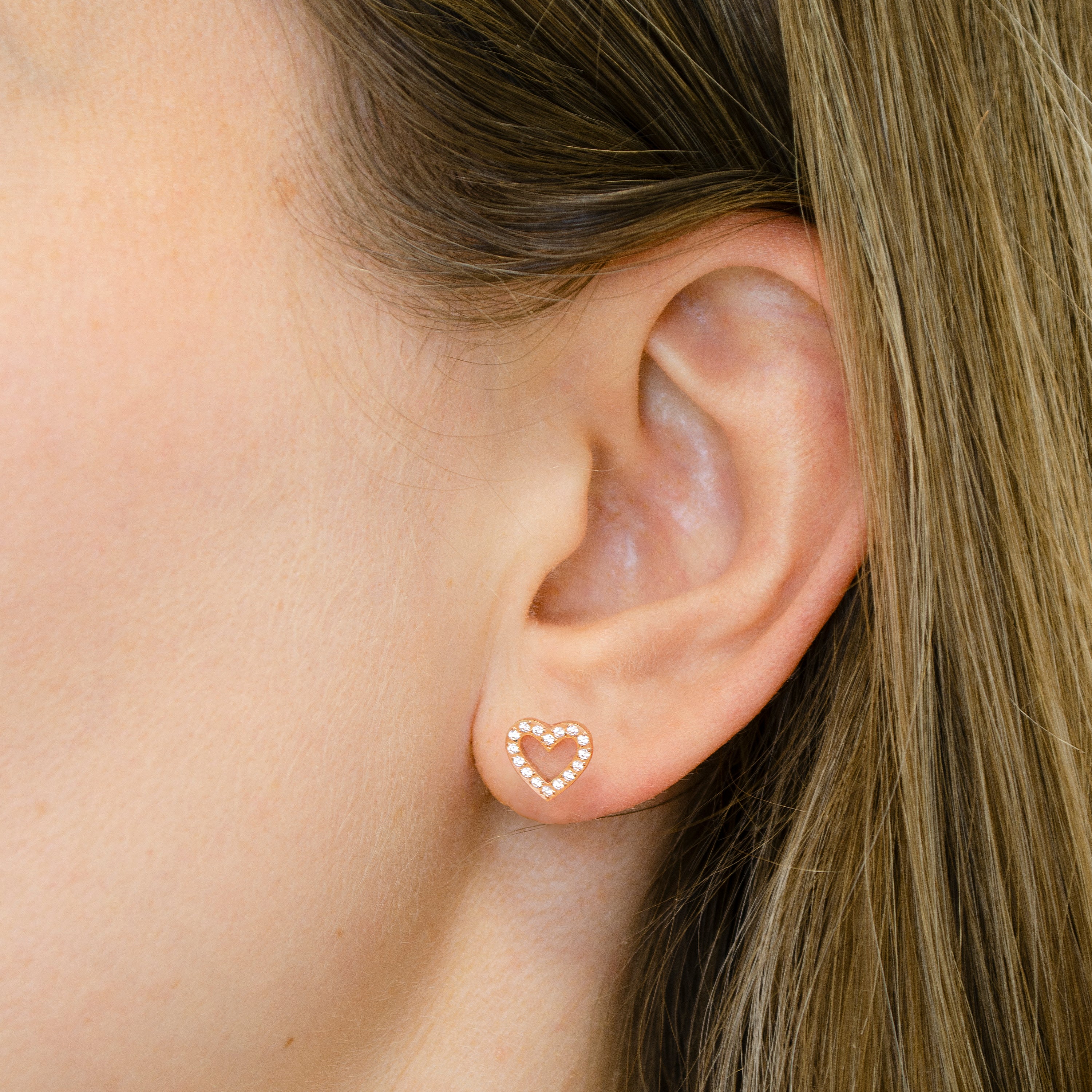 Nahaufnahme eines Ohrs mit hellbraunem Haar, das einen Tamaris Ohrstecker von Tamaris trägt, einen goldenen herzförmigen Ohrstecker, der mit kleinen Kristallen besetzt ist.