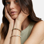 Eine Frau in einem ärmellosen Oberteil stützt ihr Gesicht in die Hände und trägt zwei Armbänder Tamaris Beads - Agalmatolith Armband von Tamaris an den Handgelenken.