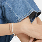Nahaufnahme einer Person in Blue Jeans, die zwei zarte Armbänder trägt - das eine ist das mit Glasperlen verzierte Armband The Pearl Beads von Tamaris, das andere besteht aus kleinen farbigen Steinen.