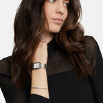 Frau mit langen braunen Haaren, die ein schwarzes Oberteil und eine Tamaris Armbanduhr - The Square Retro Strap von Tamaris trägt und nachdenklich zur Seite schaut.