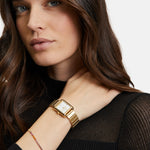 Frau mit langen braunen Haaren, schwarzem Oberteil und goldener Armbanduhr trägt das Tamaris Armband - The Rainbow Sparkling Beads im Layering-Look und schaut selbstbewusst in die Kamera.