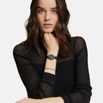 Eine Frau mit gewellten braunen Haaren in einem schwarzen Top lehnt sich an einen Tisch und betont das Tamaris Armband - The Black Sparkling Beads von Tamaris für einen schicken Lagenlook.