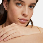 Eine Frau mit blauen Augen und einem ruhigen Gesichtsausdruck trägt Tamaris Goldschmuck und die Tamaris Creole - The Silver Collection, die einen Anhänger aus 925er Sterlingsilber mit blauen Zirkonia vor einem schlichten Hintergrund zeigt.