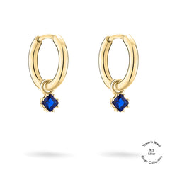 Tamaris Creole - Die Silberkollektion: Goldfarbene Creolen mit blauen Zirkonia-Anhängern, hergestellt aus 925er Sterlingsilber von Tamaris, präsentiert auf weißem Hintergrund mit einem kleinen Logo in der Ecke.