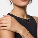Eine Frau in einem ärmellosen schwarzen Oberteil berührt ihre Schulter und trägt Goldschmuck - Ohrringe, Halskette und das Tamaris Armband - Tiny Tubes von Tamaris.