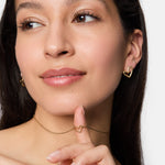 Eine Frau lächelt, trägt goldene Ohrringe in Herzform und die Tamaris Halskette - Graffiti Heart, die sanft die Kette berührt - dieses auffällige Stück von Tamaris ist das perfekte Accessoire für Frauen.