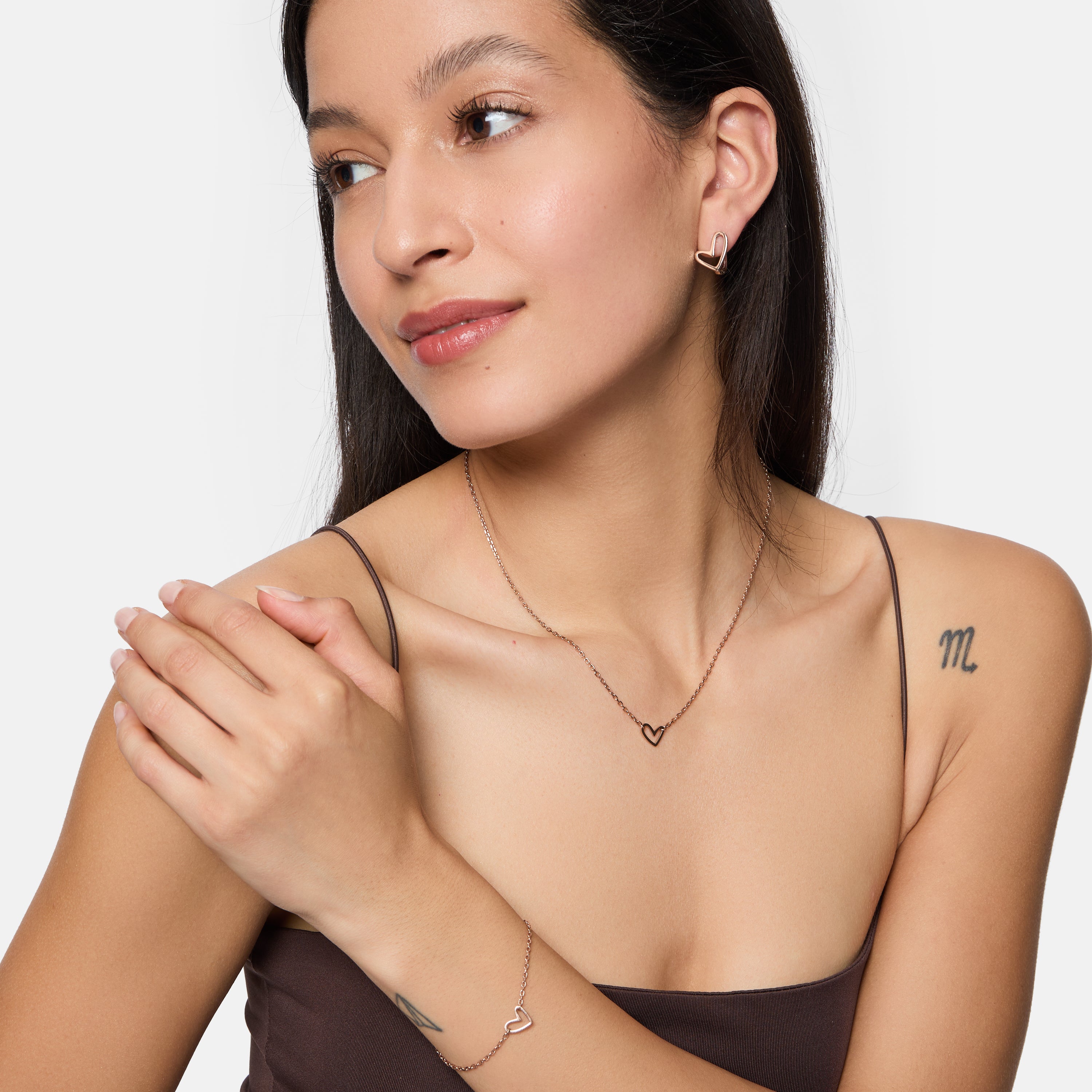 Frau trägt die Tamaris Creole - Graffiti Heart Ohrringe, Teil eines herzförmigen Schmucksets von Tamaris, bestehend aus Halskette und Armband, gepaart mit einem braunen Top und sichtbaren Tattoos.