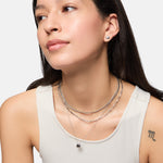 Frau in weißem Tank-Top mit mehrlagigen Silberketten und Tamaris Ohrstecker - Modern Layering von Tamaris, mit organischem Design, sowie einem "m"-Tattoo auf dem Arm.