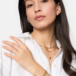 Eine Frau in einem weißen Hemd trägt Goldschmuck, darunter das Tamaris Armband - Modern Layering von Tamaris, Gliederketten, Armbänder, Ohrringe und hat ein sichtbares Handgelenk-Tattoo.