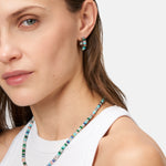 Eine Frau, die die Tamaris Halskette - All About Beads und passende Ohrringe zu einem weißen Tanktop trägt, schaut in die Kamera.