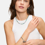 Frau in einem weißen, ärmellosen Oberteil, mit passender Perlenkette und Ohrringen, ausgestattet mit dem Tamaris Armband - All About Beads by Tamaris, und mit einem sanften Lächeln posierend.
