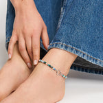 Eine Trägerin der Tamaris Fußkette - All About Beads by Tamaris mit bunten Perlen und einem silbernen Ring, bekleidet mit Blue Jeans, berührt sanft ihren Knöchel.