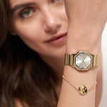 Eine Frau trägt ihre goldene Armbanduhr und das Tamaris Armband - Little Twinkle von Tamaris mit einem runden Anhänger mit funkelnden Zirkoniasteinen in der Nähe ihres Gesichts.