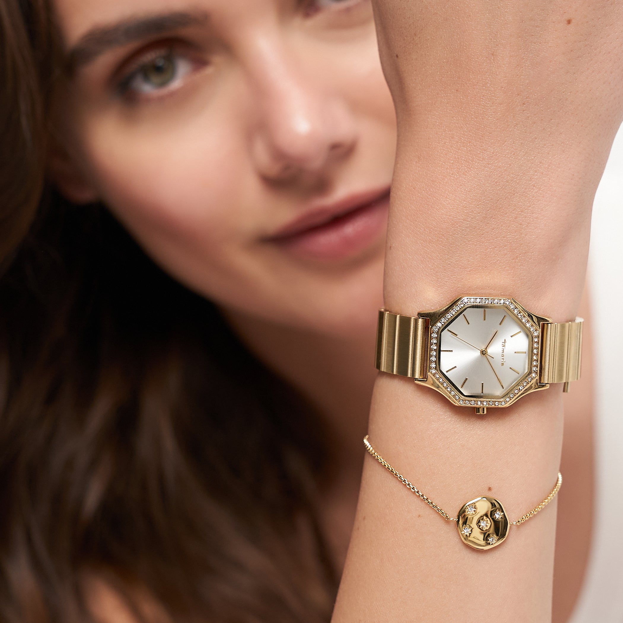 Eine Frau trägt ihre goldene Armbanduhr und das Tamaris Armband - Little Twinkle von Tamaris mit einem runden Anhänger mit funkelnden Zirkoniasteinen in der Nähe ihres Gesichts.