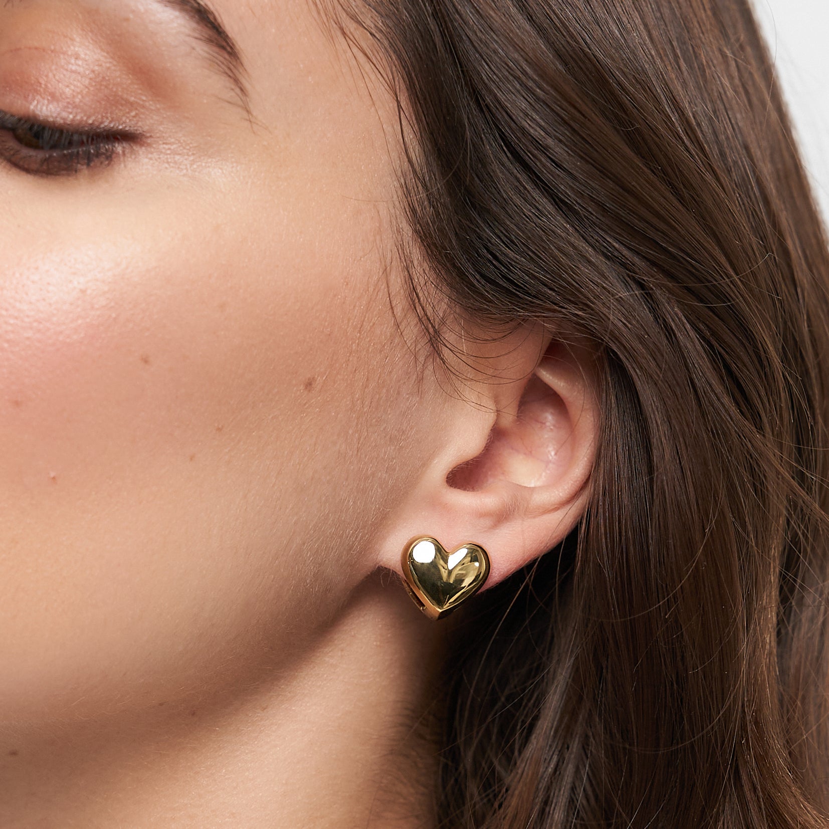 Nahaufnahme eines Frauenohrs, das den Tamaris Ohrstecker - Double Heart von Tamaris trägt. Das goldglänzende Doppelherz ist mit braunen Haaren besetzt - ein elegantes Accessoire für jede Gelegenheit.