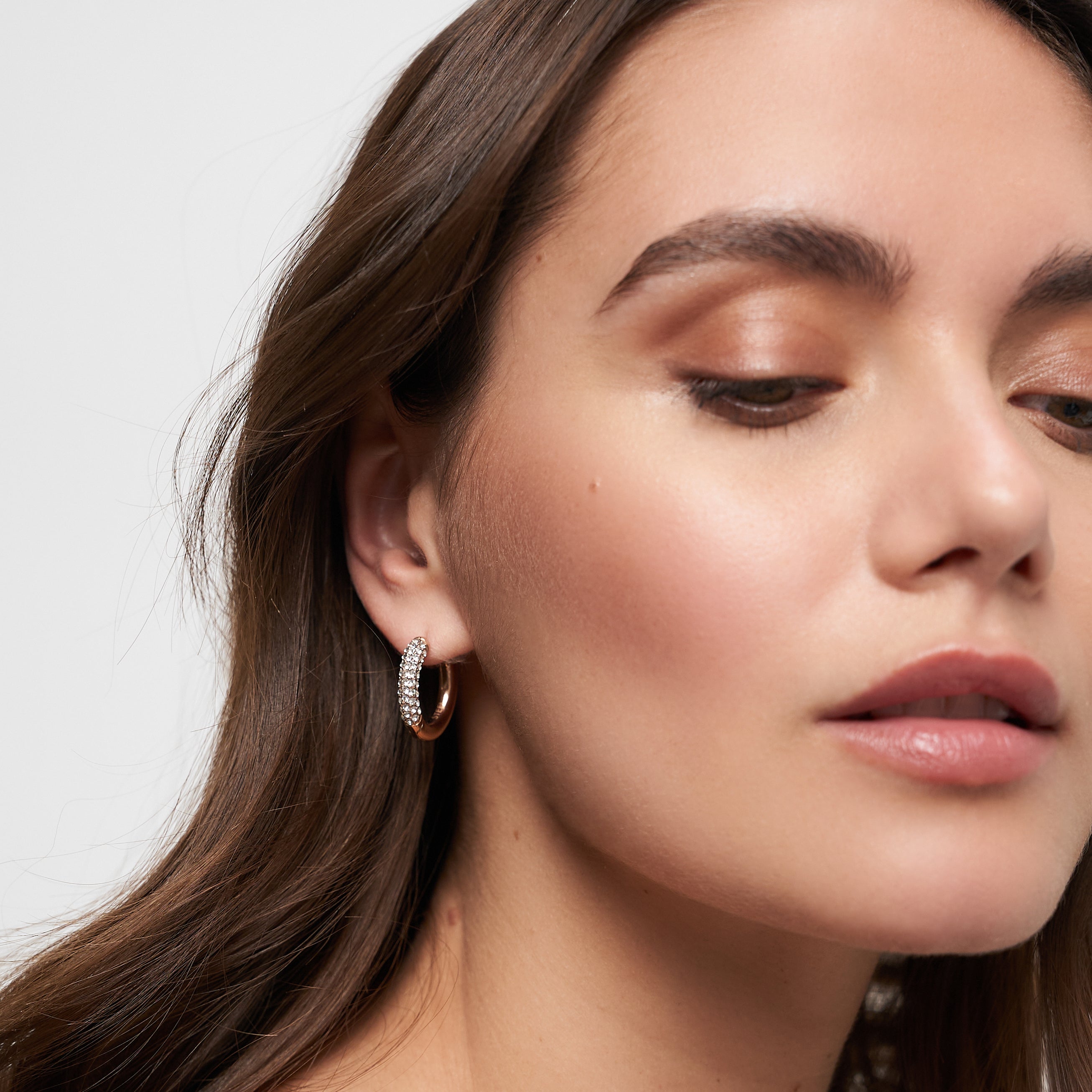 Eine Frau mit gewelltem braunem Haar, die Tamaris Creole - Glitter Donuts silberne Creolen trägt, die mit funkelnden Steinen verziert sind, sieht mit natürlichem Make-up leicht nach unten.