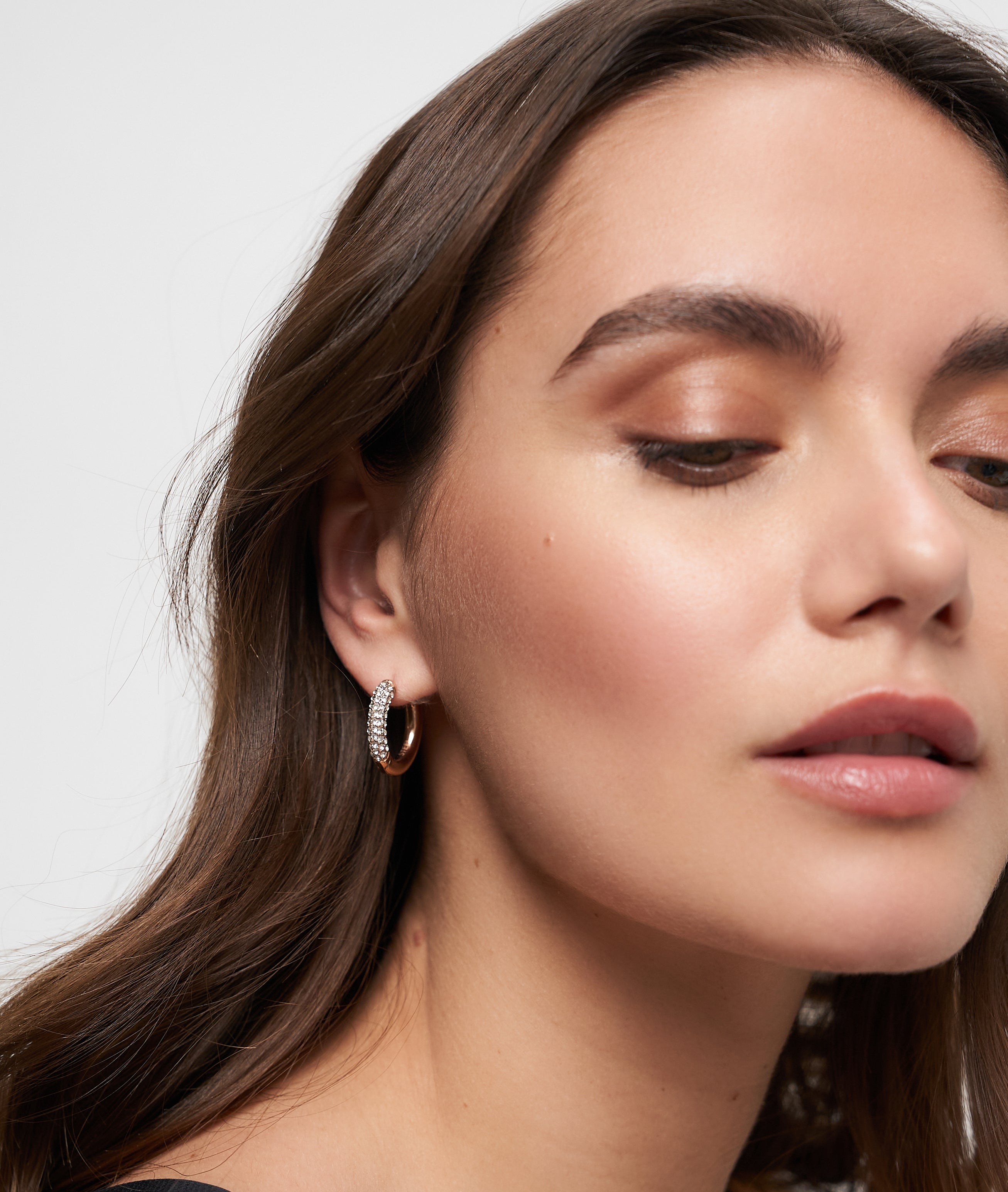 Eine Frau mit gewelltem braunem Haar, die Tamaris Creole - Glitter Donuts silberne Creolen trägt, die mit funkelnden Steinen verziert sind, sieht mit natürlichem Make-up leicht nach unten.