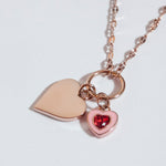 Die Tamaris Halskette - Charm Series ist eleganter Schmuck: eine roségoldene Kette mit zwei Herzanhängern von Tamaris, einer schlicht, der andere mit rotem Edelstein geschmückt.