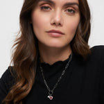 Eine Frau mit gewelltem braunem Haar trägt ein schwarzes Oberteil und eine Tamaris Charms Bundle - Charm Series Halskette mit einem rosa Herzanhänger der Marke Tamaris.