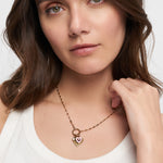 Eine Frau in einem weißen Tank-Top trägt eine Tamaris Charms Bundle-Halskette aus der Charm-Serie und hält die goldene Kette am Hals.