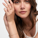 Frau mit langen braunen Haaren trägt goldenen Schmuck, darunter einen LOVE-Anhänger und ein Tamaris-Armband aus der Silver Love Collection von Tamaris, gepaart mit einem weißen Oberteil, und stützt ihre Hand auf ihr Gesicht.