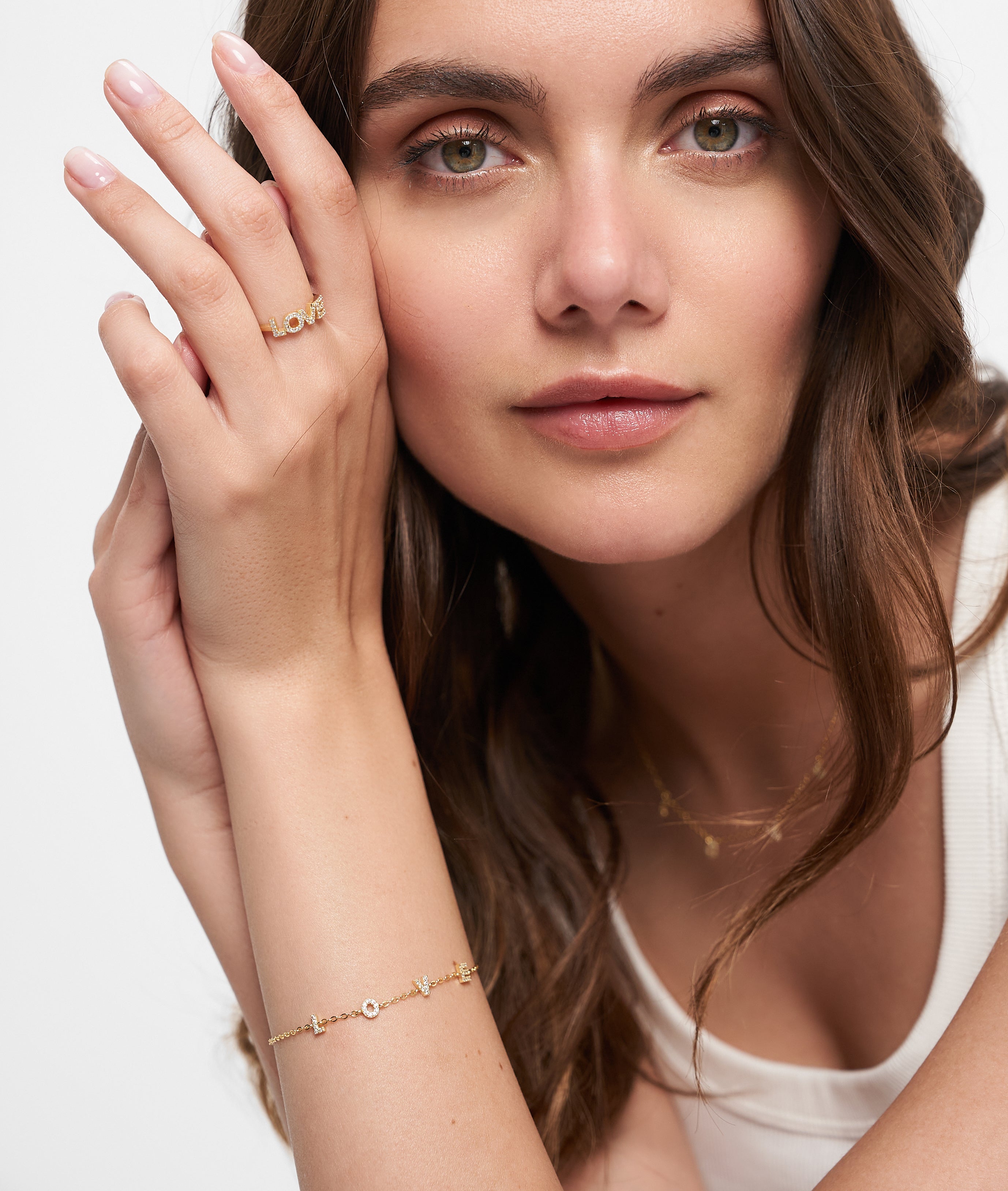 Frau mit langen braunen Haaren trägt goldenen Schmuck, darunter einen LOVE-Anhänger und ein Tamaris-Armband aus der Silver Love Collection von Tamaris, gepaart mit einem weißen Oberteil, und stützt ihre Hand auf ihr Gesicht.