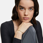 Frau mit blauen Augen und dunklem Haar stützt ihr Kinn auf die Hand. Sie trägt Goldschmuck - eine Schlangenkette und ein elegantes Armband - und ein schwarzes Tamaris Set - The Layer Top.