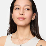 Frau mit langen braunen Haaren, trägt ein weißes Tanktop, Ohrringe, Damen Schmuck, eine Tamaris Halskette - Modern Layering von Tamaris und ein m-Tattoo auf ihrer Schulter.
