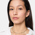 Eine Frau mit langen dunklen Haaren trägt ein weißes Hemd, goldene Ohrringe und die Tamaris Halskette - Modern Layering von Tamaris und schaut in die Kamera.