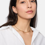 Frau in weißem Hemd mit Tamaris Halskette - Modern Layering und kleinen goldenen Ohrringen. Ihr glattes dunkles Haar vervollständigt einen eleganten Look mit Damen Schmuck und dezent gestufter Schlangenkette.