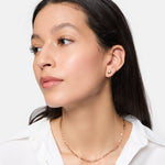 Frau mit langen dunklen Haaren trägt ein weißes Hemd und die Tamaris Halskette - Modern Layering von Tamaris, gepaart mit kleinen Ohrsteckern, mit Blick zur Seite.