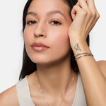 Frau mit langen dunklen Haaren in einem Tanktop, die das Tamaris Armband - Modern Layering von Tamaris und eine Halskette trägt, zeigt ein Papierflugzeug-Tattoo auf ihrem Handgelenk.