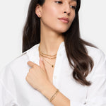 Frau mit weißem Hemd trägt Goldschmuck: eine Schlangenkette, mehrere Halsketten, Tamaris Armband - Modern Layering Armbänder von Tamaris, Ohrringe und einen Ring.