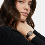 Frau mit langen braunen Haaren in einem schwarzen Pullover trägt die Tamaris Armbanduhr - The Square Black und schaut in die Kamera.