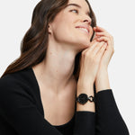 Frau in schwarzem Top, lächelnd, trägt die Tamaris Armbanduhr - The Black Glamorous mit Edelstahlband, die Hände vor dem Gesicht verschränkt.