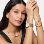 Eine Frau mit langen dunklen Haaren posiert und trägt eine goldene Uhr, eine Kettenhalskette und das Tamaris Armband - Modern Pearl Fusion von Tamaris; ihre Tätowierungen sind deutlich sichtbar.