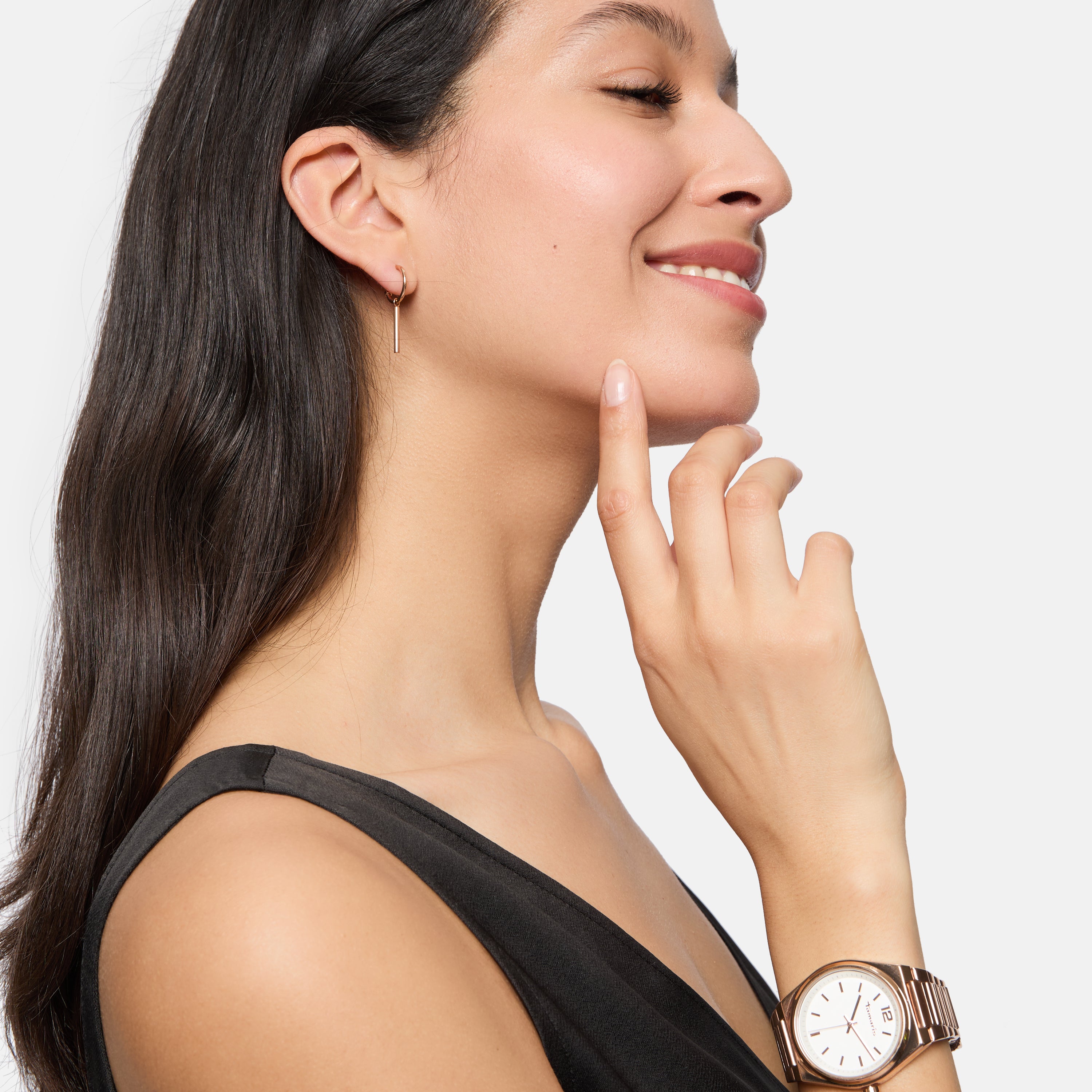 Eine Frau in einem ärmellosen Oberteil lächelt im Profil und berührt ihr Kinn. Sie trägt die Tamaris Creole - Modern Chain Ohrringe und eine Tamaris-Uhr - ein Beispiel für minimalistisches Design.