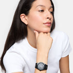 Eine Frau im weißen Hemd stützt gedankenverloren ihr Kinn auf die Hand und trägt die Tamaris Armbanduhr - The Round Basic Essential, die zeitlose Eleganz ausstrahlt.